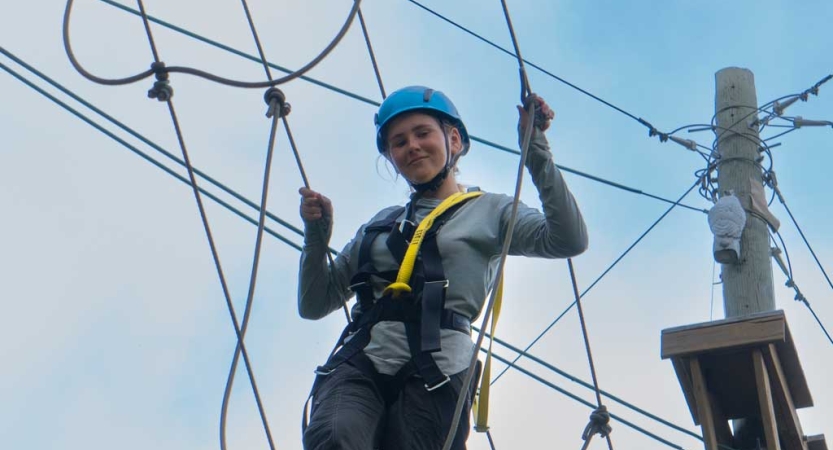 A student wearing safety gear is secured by ropes as they navigate a high ropes course. 